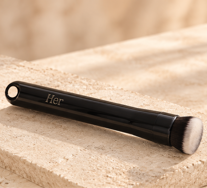 The black Her Cosmetique electric foundation brush with its gold Her handle branding, shown in full on a stone surface